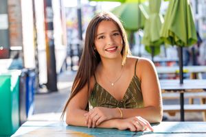 Young woman with long brown hair smiles at outdoor cafe table, wearing olive green top on sunny day.