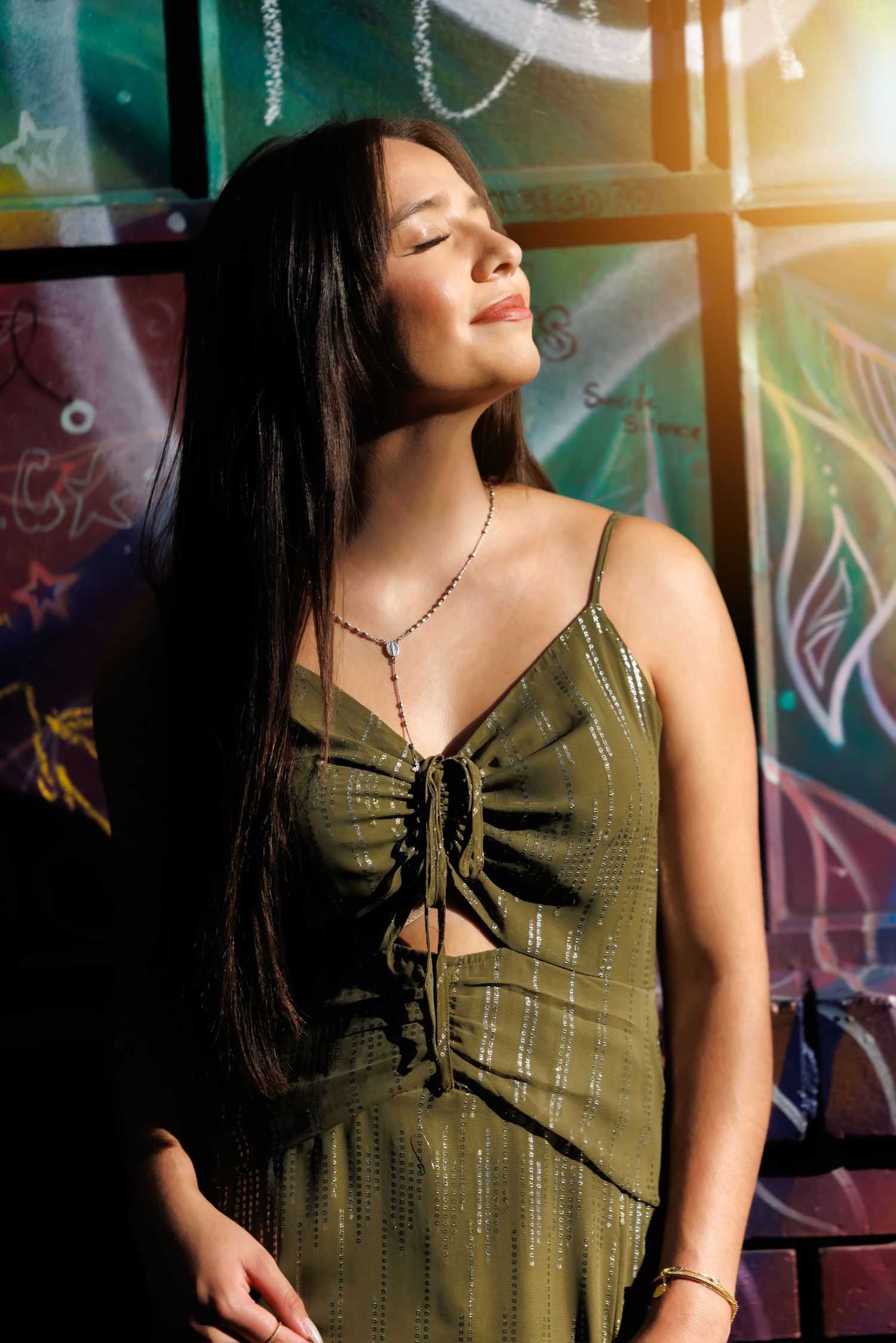 Young woman in olive green dress with eyes closed, basking in sunlight against colorful graffiti wall.