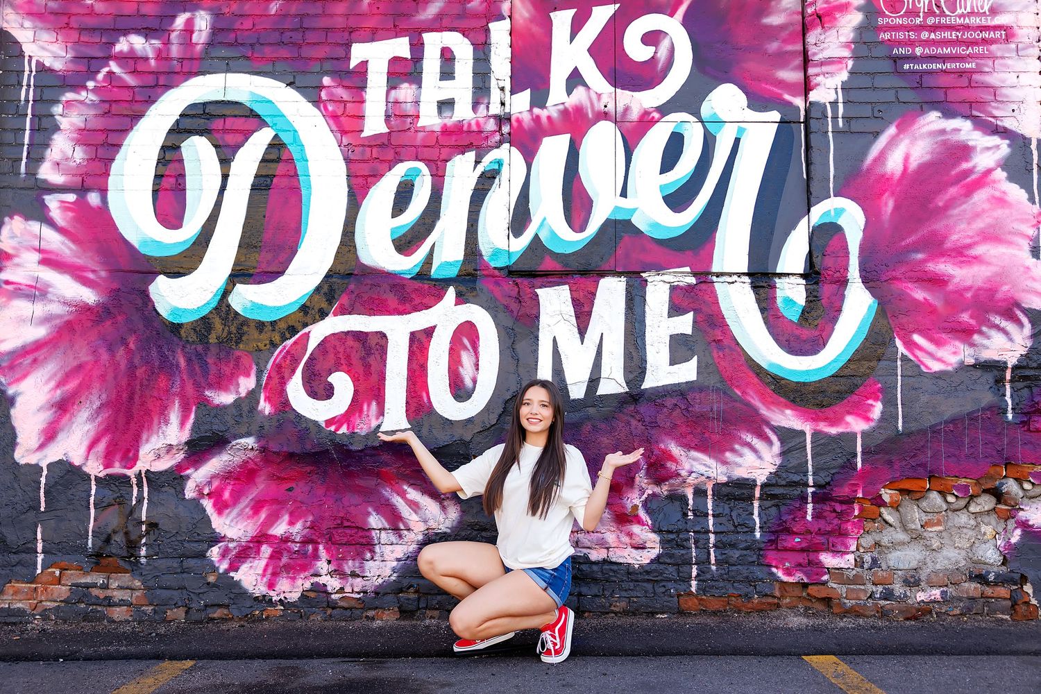 Woman posing in front of a colorful Talk Denver To Me mural with pink floral graffiti art in Denver, Colorado.