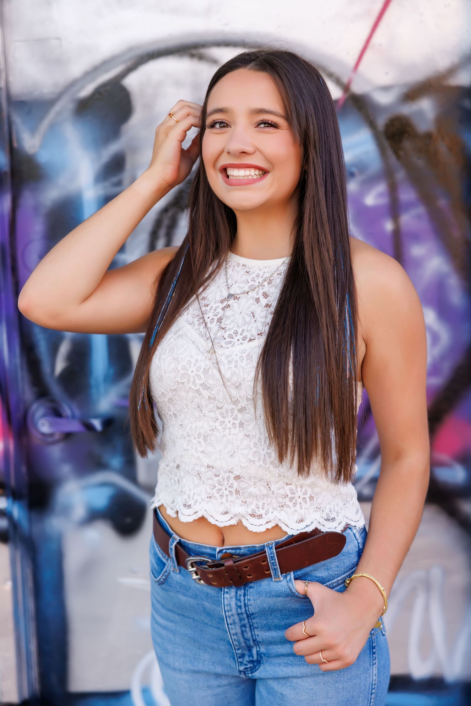 Smiling young woman in white lace crop top and jeans posing in front of colorful graffiti wall.