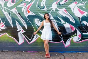 Young woman in white dress posing confidently in front of colorful graffiti wall mural outdoors.