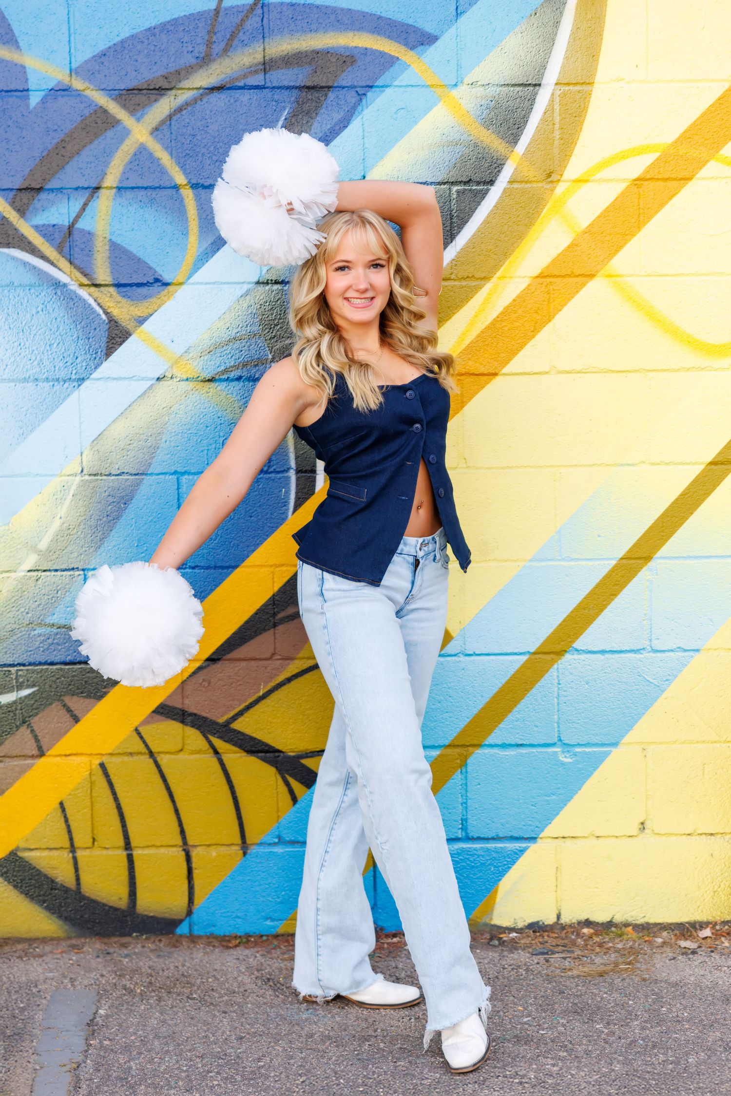 Cheerleader poses in front of vibrant blue and yellow graffiti wall wearing denim and white pom poms.