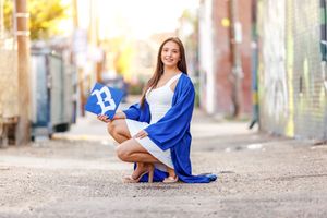 A young woman in a blue graduation gown kneels outdoors holding a letter B, celebrating her academic achievement.