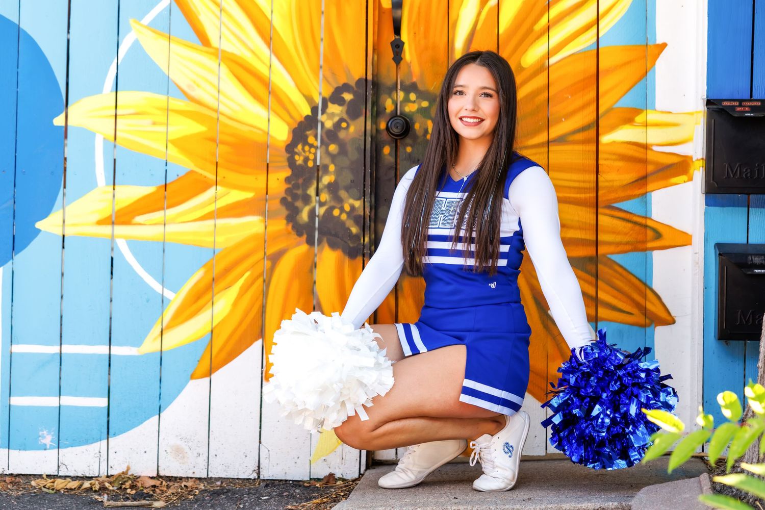 Cheerleader in blue and white uniform poses with pom-poms in front of a vibrant sunflower mural.