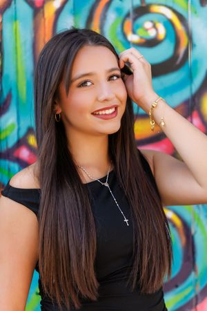 Young Latina woman with long dark hair smiling, wearing black top and cross necklace, posing against colorful graffiti wall.