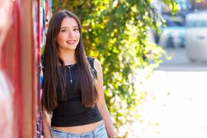 Smiling young woman with long dark hair wearing a black crop top and jeans, posing outdoors near greenery.