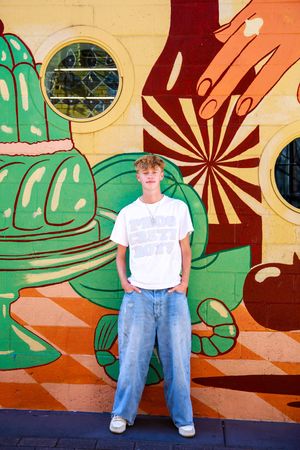 Person in white t-shirt and jeans poses against vibrant tropical mural with monstera leaves and geometric patterns.