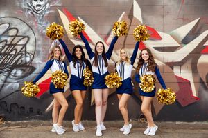 Group of cheerleaders in navy and white uniforms with gold pom-poms posing together against graffiti wall backdrop.