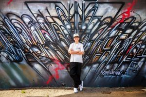 A person stands confidently against a vibrant blue and black graffiti wall with intricate designs and red accents.