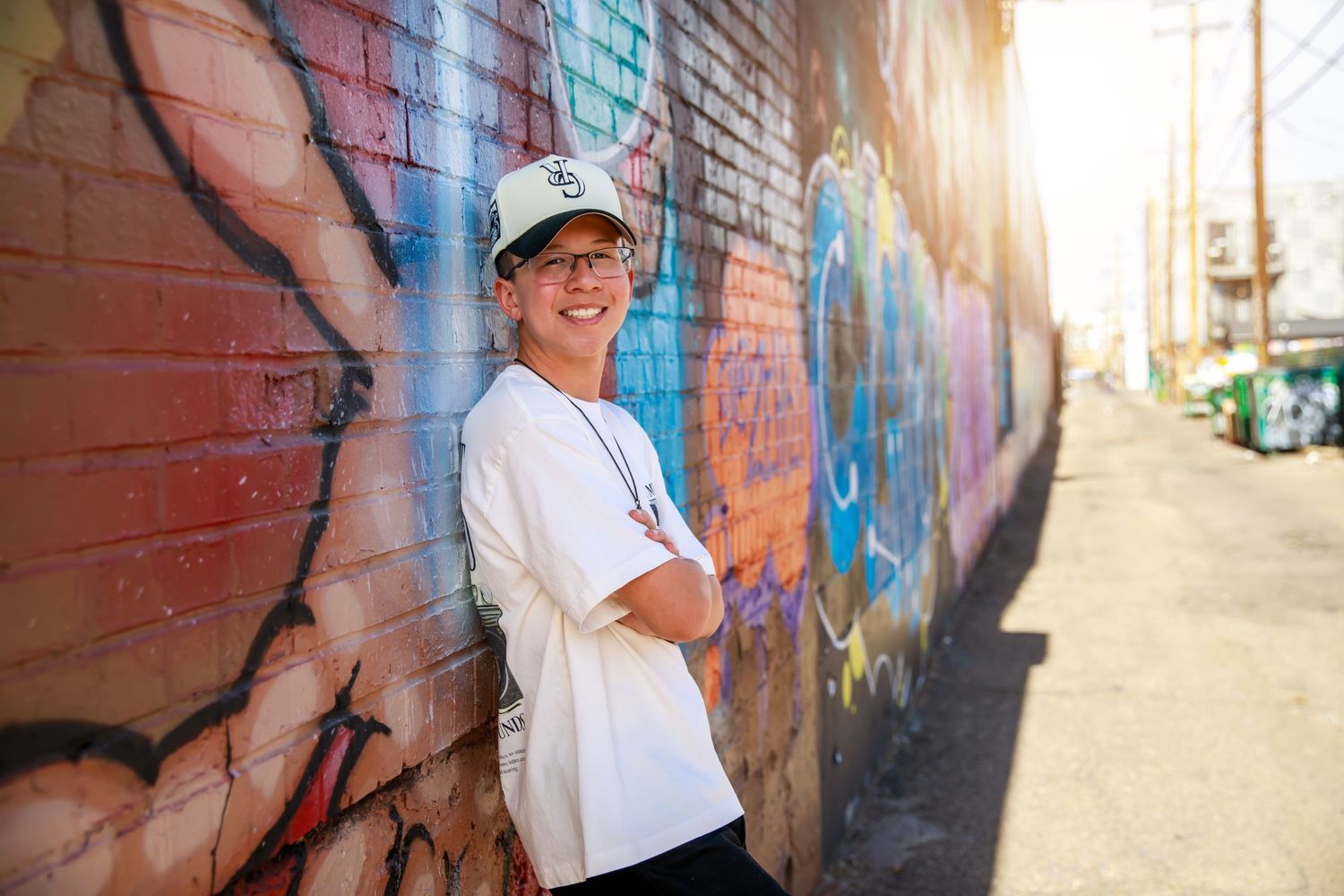 A person wearing a white t-shirt and baseball cap stands smiling against a colorful graffiti wall in sunlight.