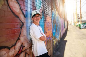 A person wearing a white t-shirt and baseball cap stands smiling against a colorful graffiti wall in sunlight.