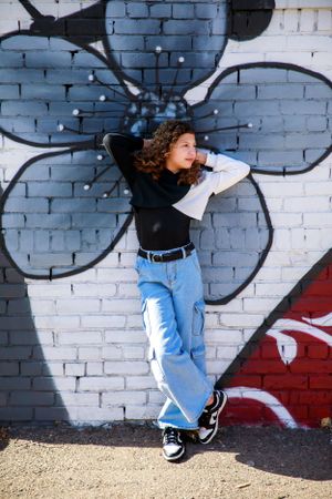 A person in light jeans and black top poses casually against a brick wall with graffiti featuring a large black flower design.