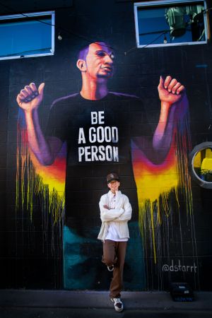 Street art mural showing a large figure wearing a 'BE A GOOD PERSON' shirt with colorful dripping paint effect on dark wall.