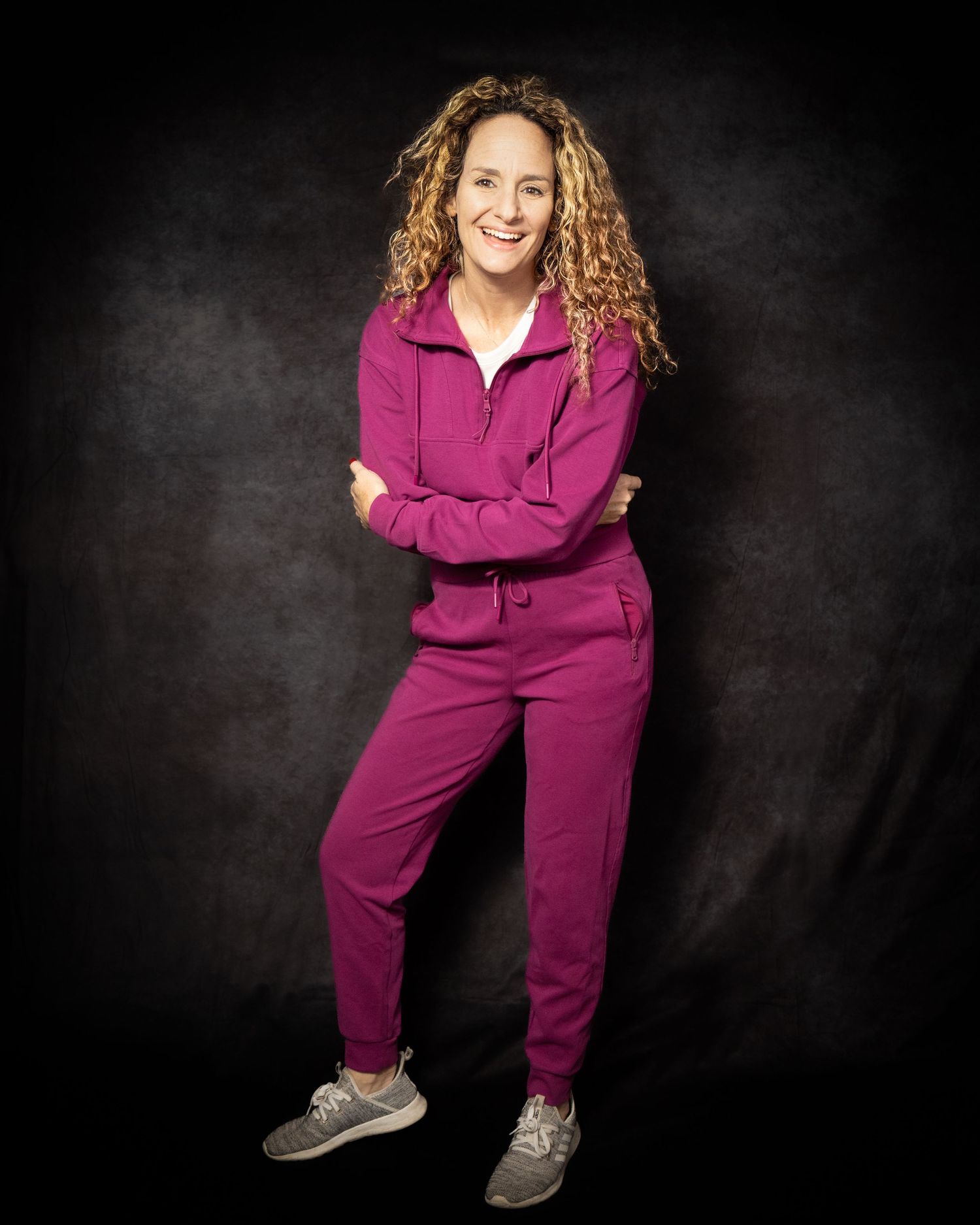 A person wearing a magenta tracksuit and sneakers stands with crossed arms against a dark background.