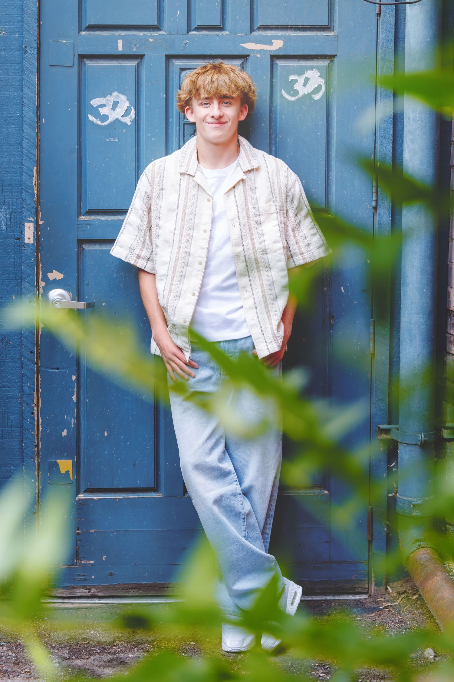 Young man with curly red hair in striped shirt and jeans leaning against a blue door surrounded by greenery.