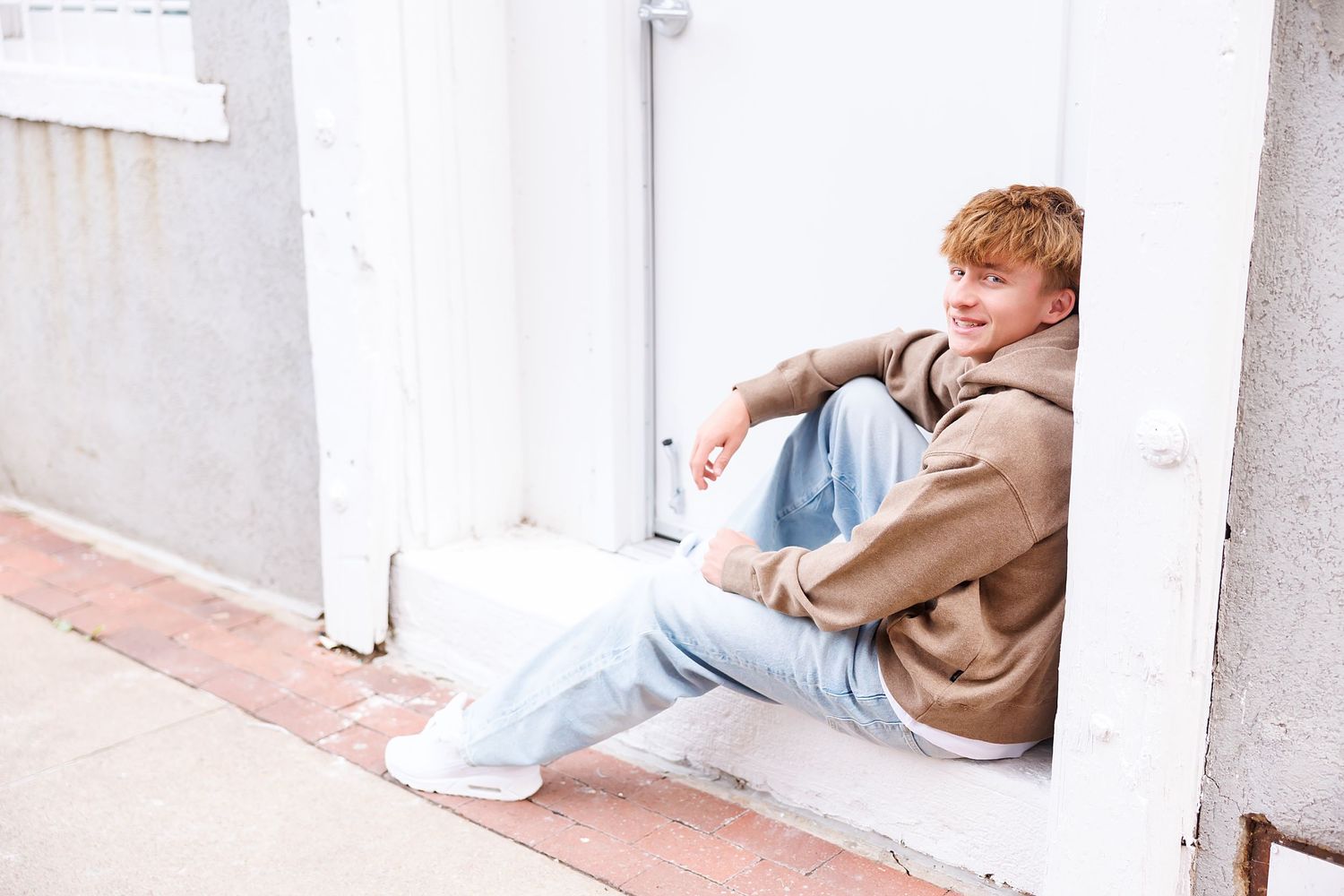 A young redhead teen boy sits relaxed in a doorway wearing a beige hoodie and light blue jeans outdoors.