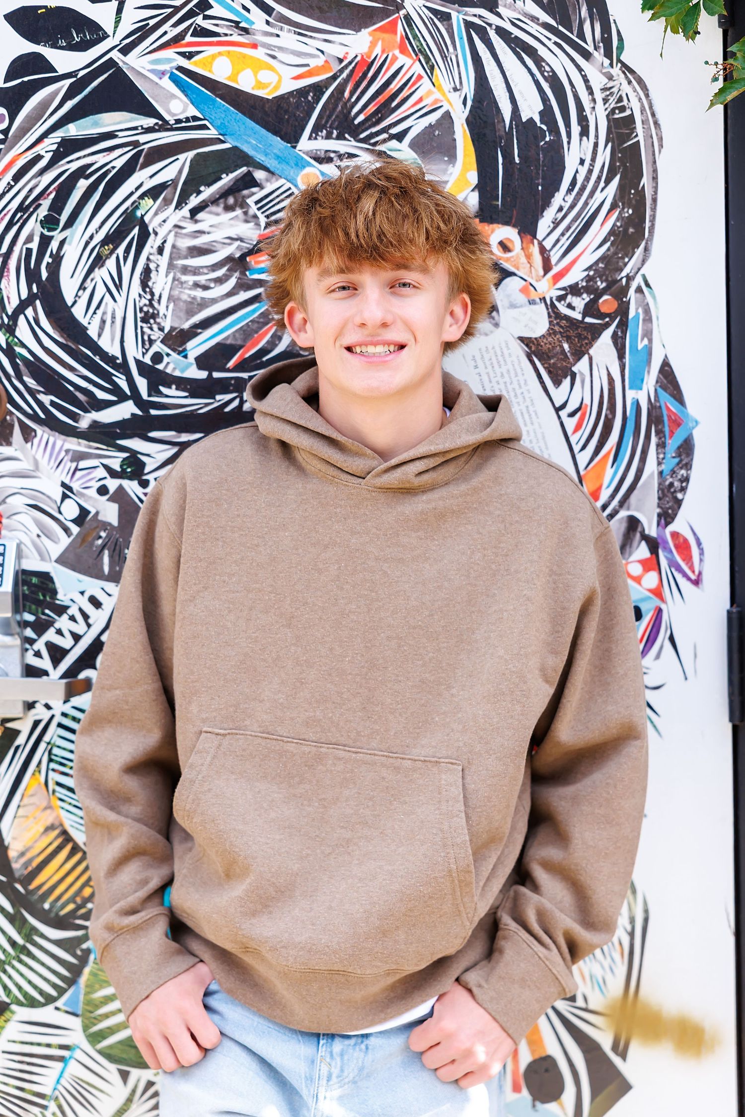 Smiling teenage boy with red hair wearing a tan hoodie, posing in front of colorful graffiti wall art.