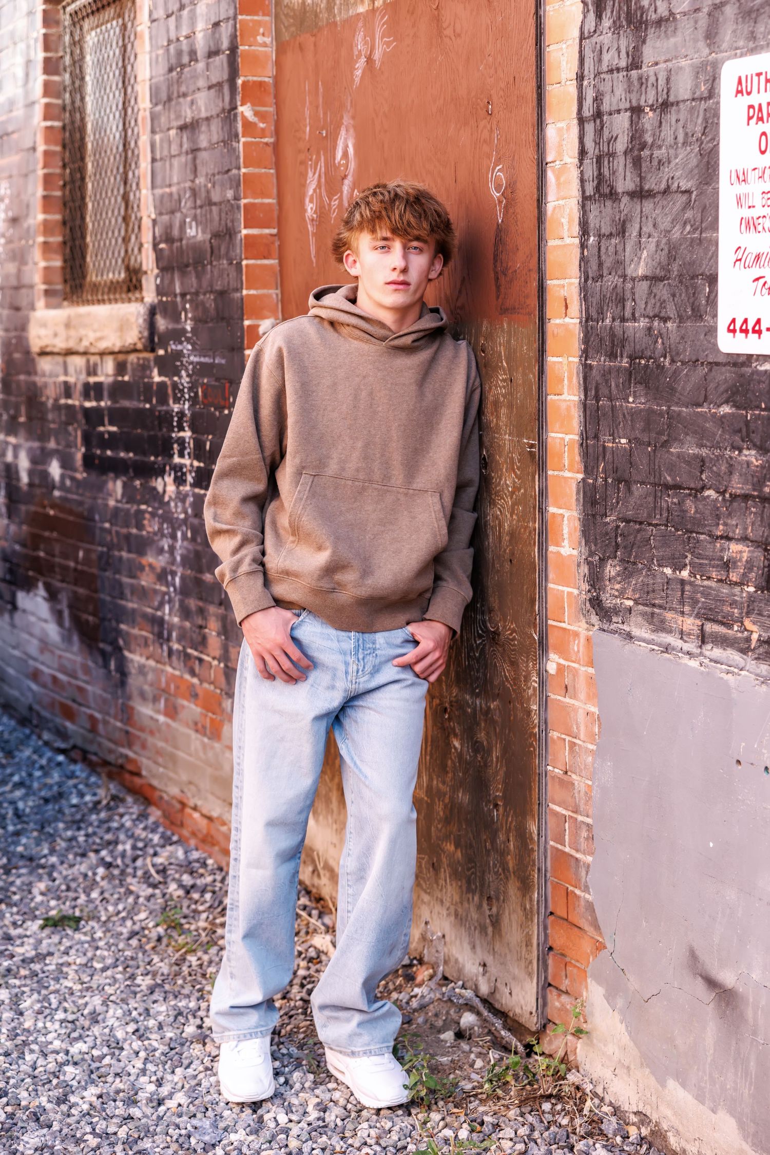 A teenage boy in a brown hoodie and light jeans leans against a weathered brick wall on a gravel surface.