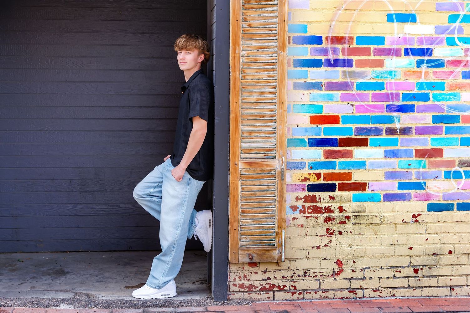 Teen boy in black shirt and jeans leans against colorful brick wall mural near a garage door.
