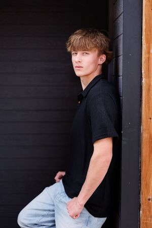 Young man in black polo shirt and jeans leaning against dark wall, looking away confidently.