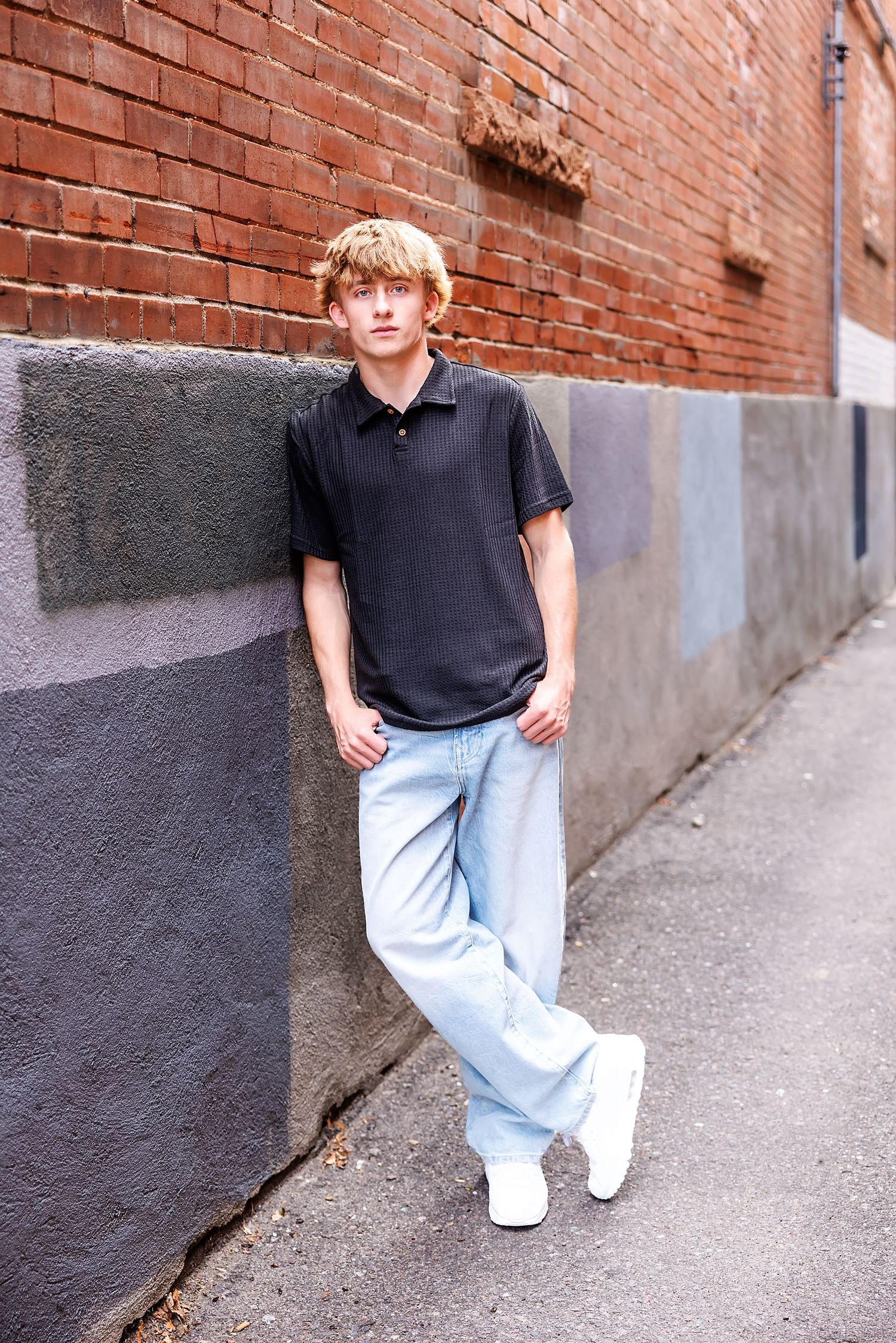 Teen boy in black polo and light jeans leans against a brick wall in an urban alley wearing white shoes.