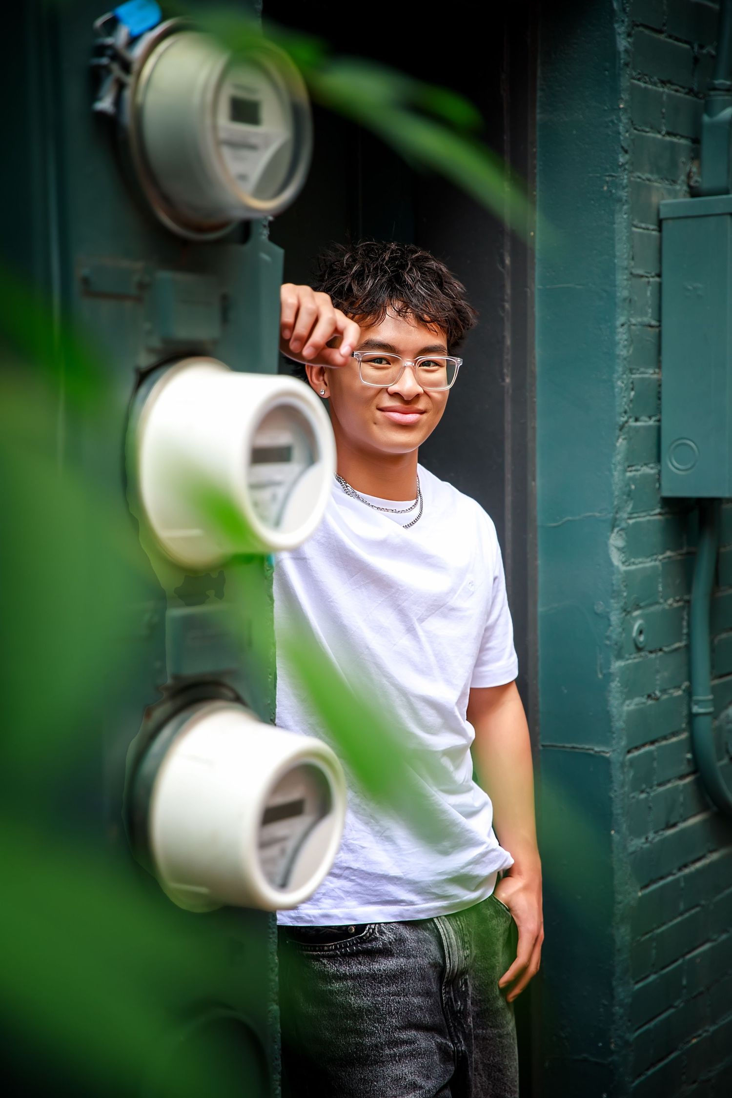 A young person in a white t-shirt and glasses poses against a teal wall with blurred foreground elements.