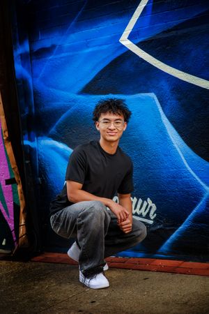 A person in a black t-shirt poses against a vibrant blue graffiti wall in an urban setting.