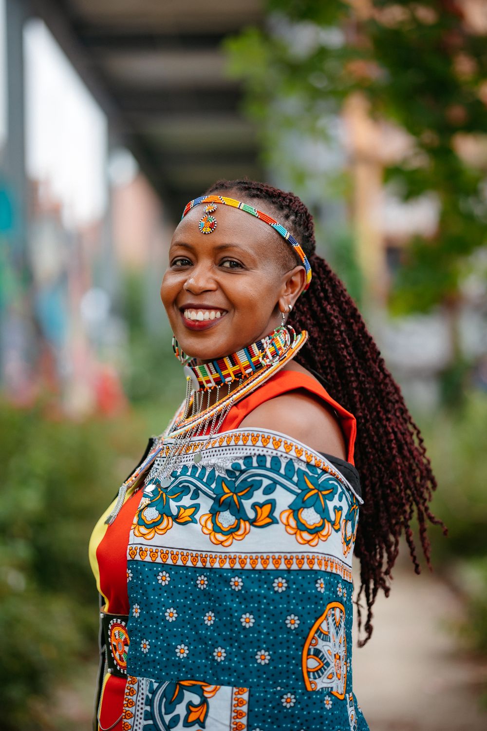 Woman in vibrant African print dress with beaded necklace and braided hair.
