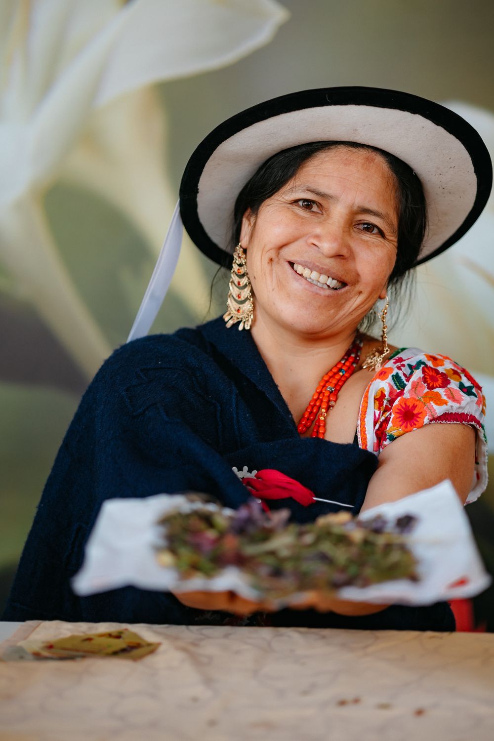 Woman in traditional Andean dress and hat presenting food at a table.