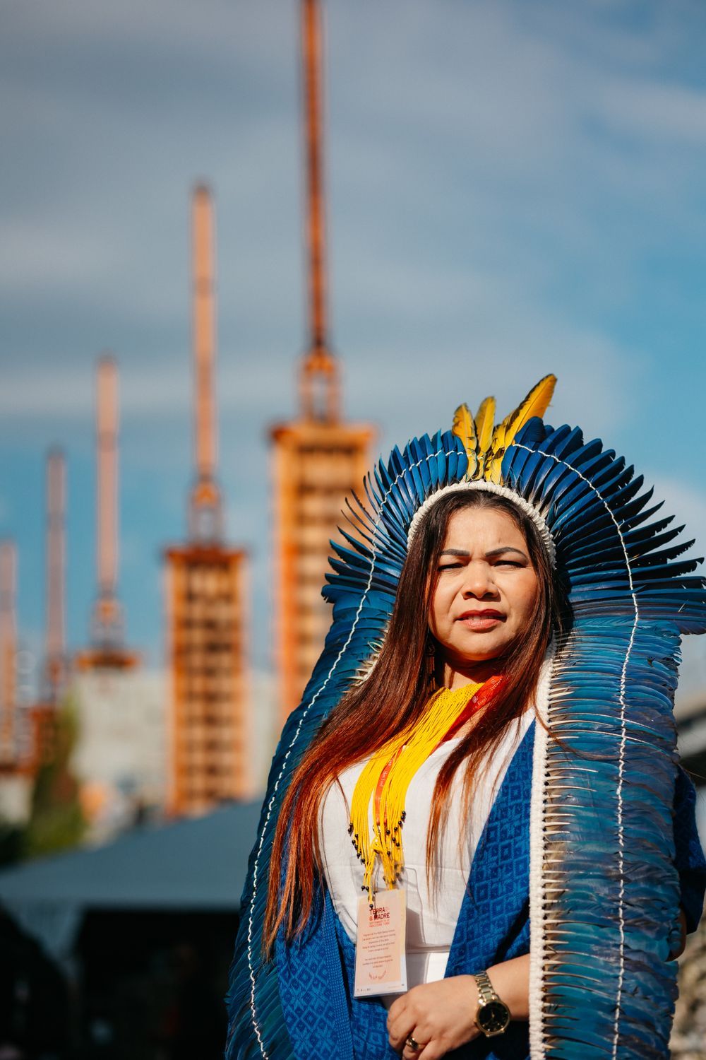Person wearing ceremonial blue feathered headdress against industrial backdrop.