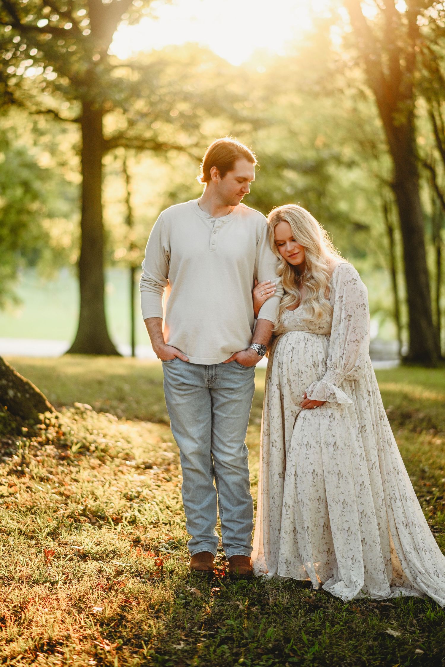 Romantic maternity photo of couple in white dress and casual wear during golden hour in park.