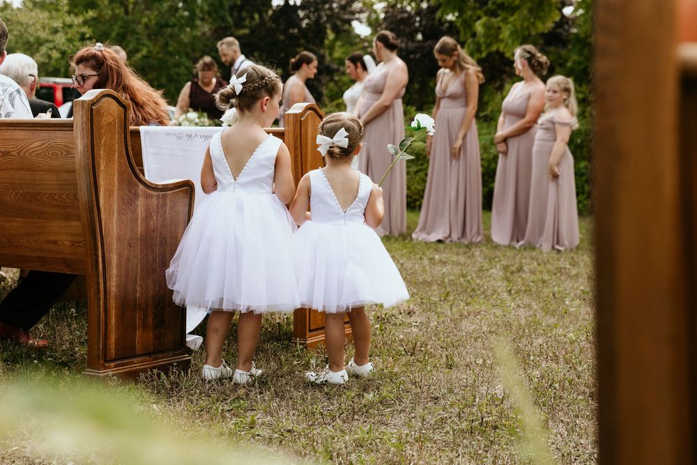 An outdoor country wedding ceremony setup features vintage wooden church pews arranged on grass under tall trees.
