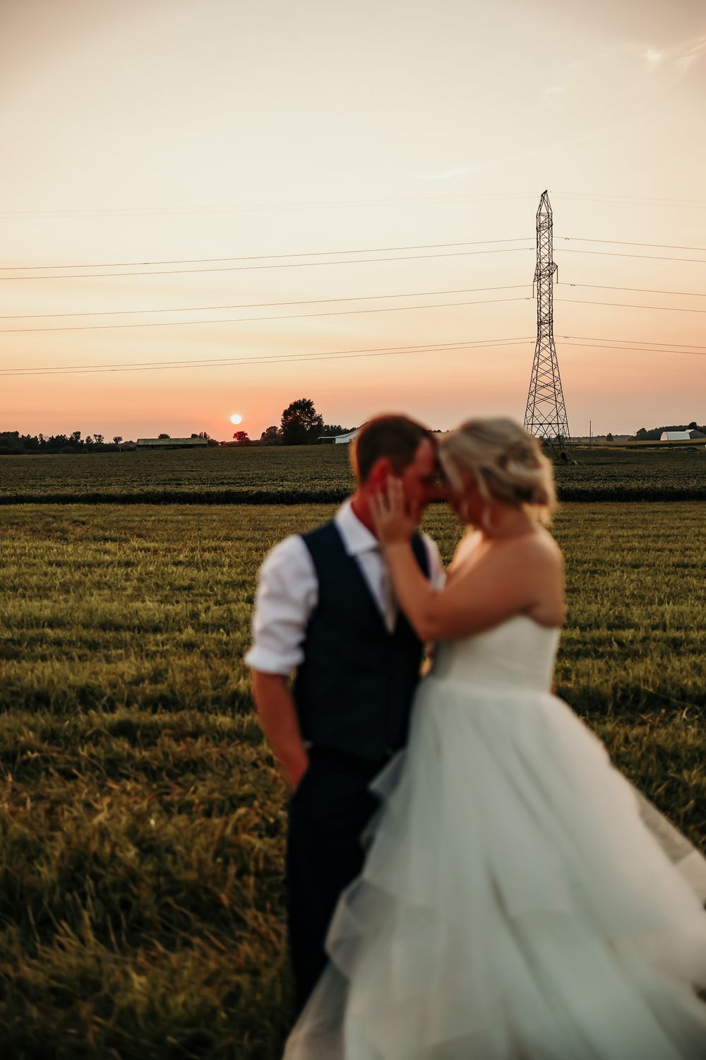 A romantic couple in formal wedding attire embraces in a sunset-lit field with power lines in the background.