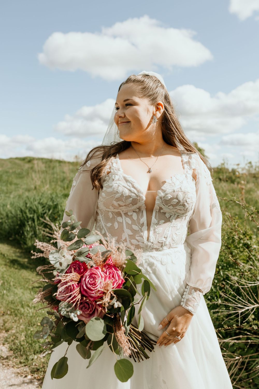A bride in a stunning lace wedding gown with long sleeves holds a vibrant pink and burgundy bouquet in a grassy field.