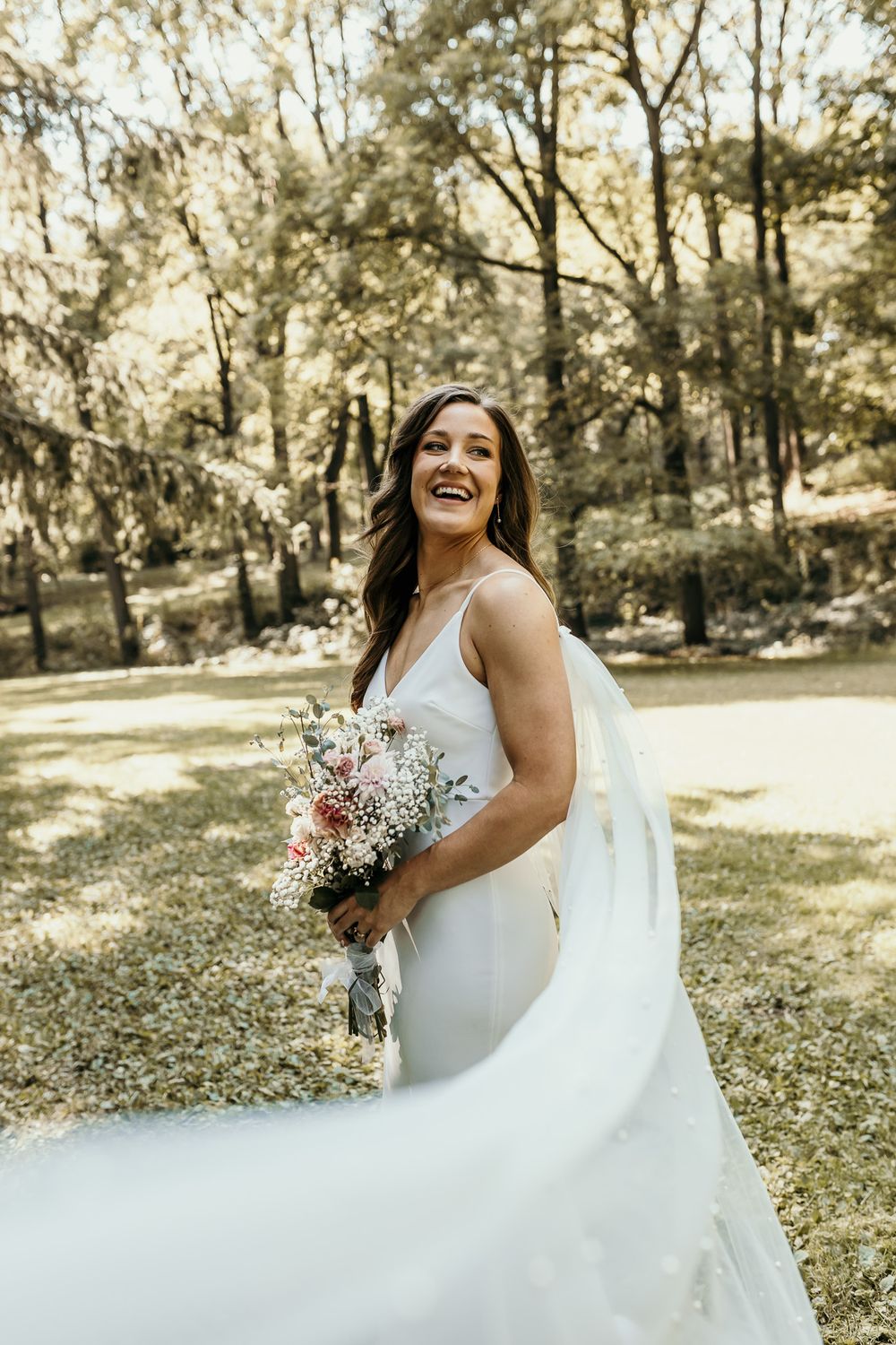 Joyful bride laughing while holding bouquet in sunlit garden setting.