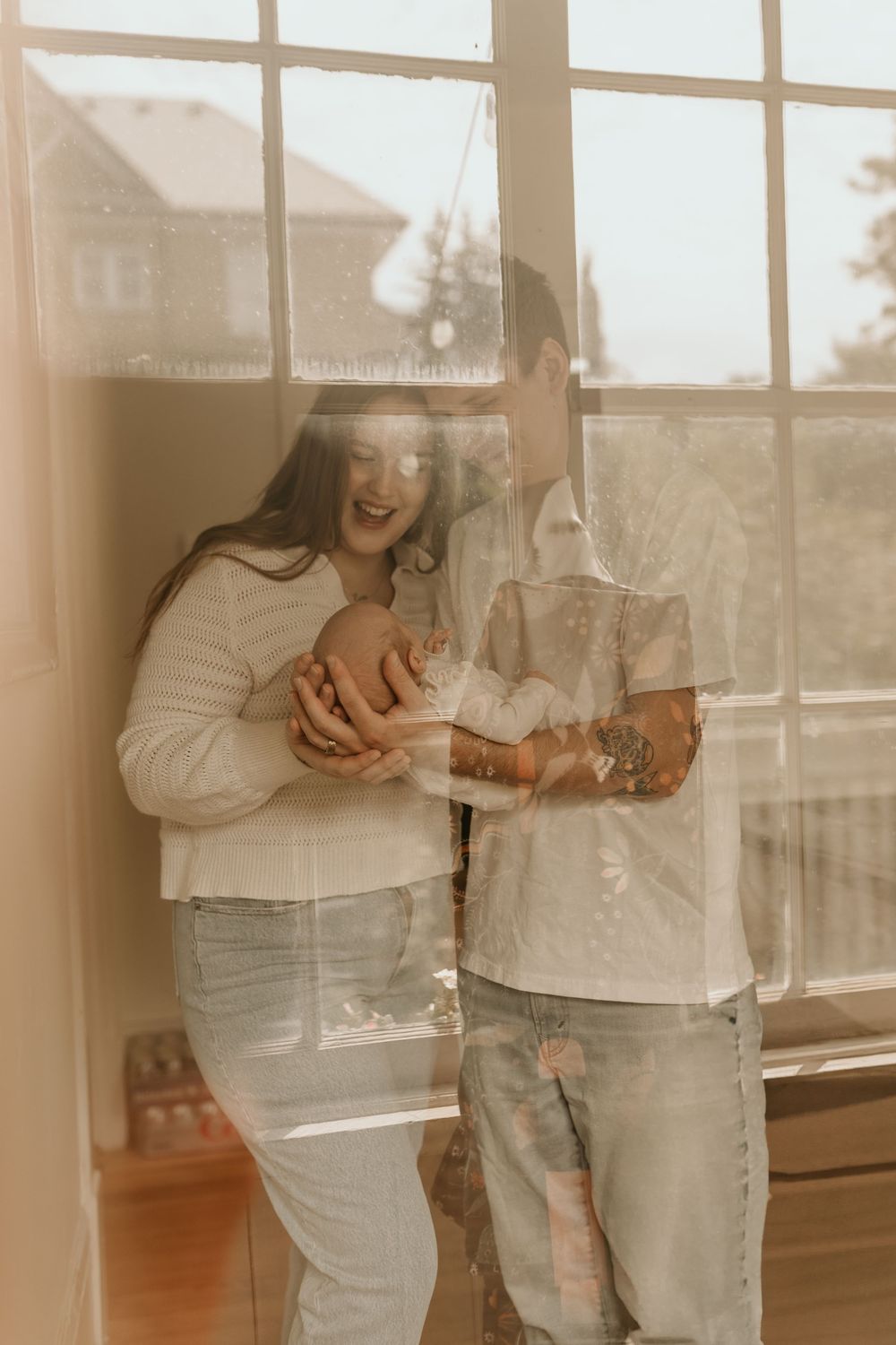 Couple wearing matching white sweaters holds coffee mugs while standing by bright sunlit windows.