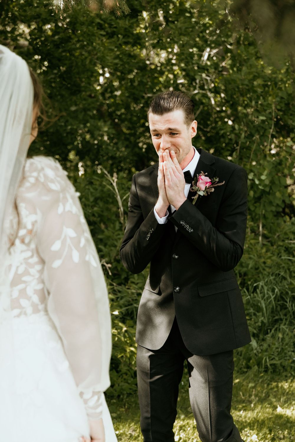 Emotional groom in black suit covers his face while seeing bride during first look wedding moment in garden setting.