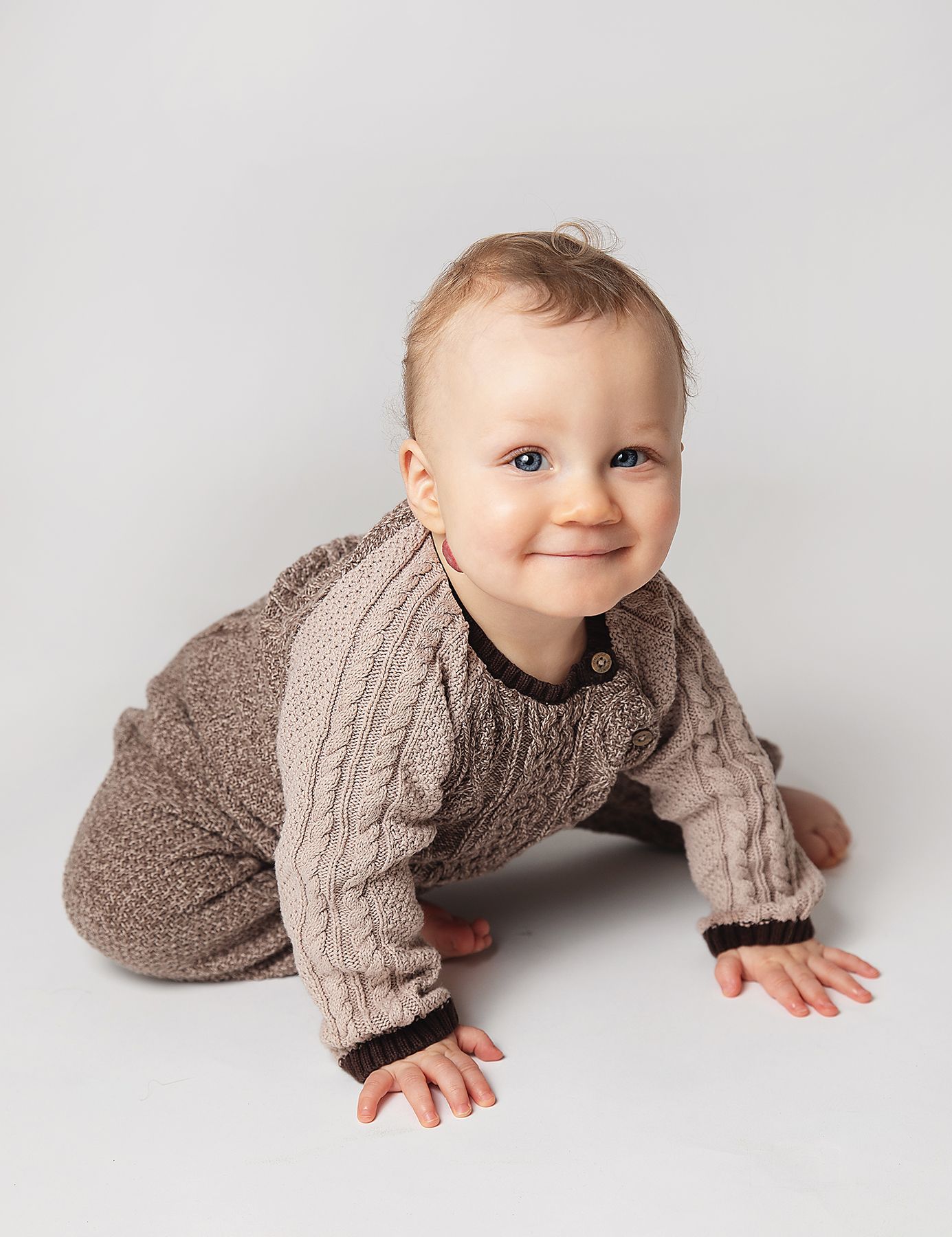 Small child wearing a cozy beige cable knit sweater crawls on white background. During a photoshoot in Bexley