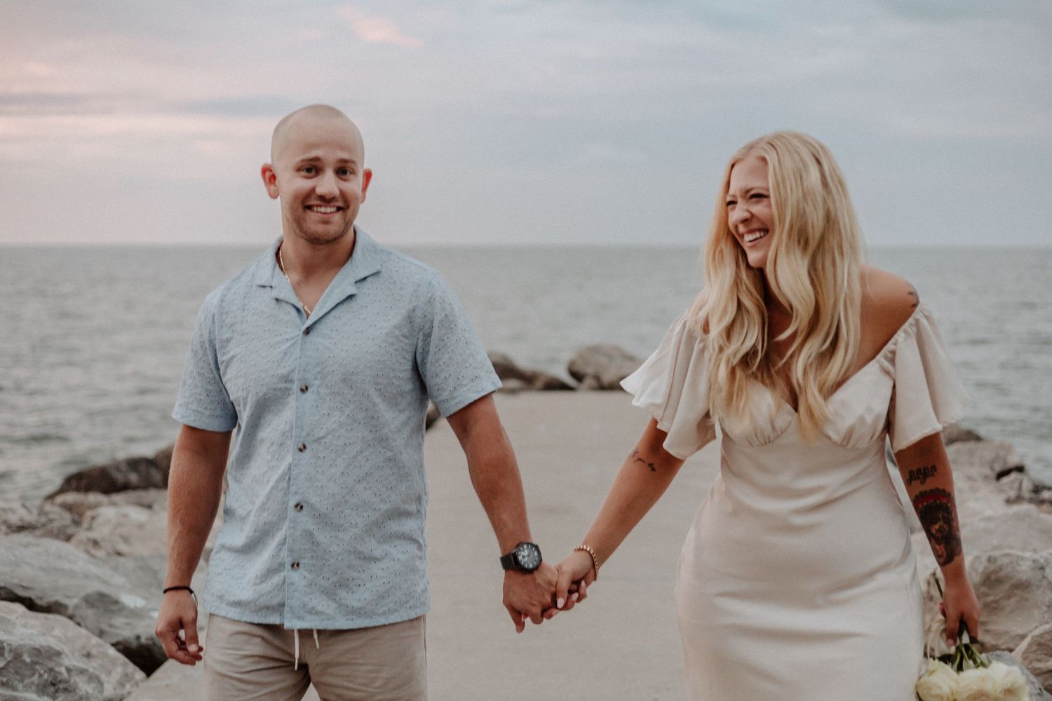 Couple holding hands on rocky beach during sunset by the ocean.
