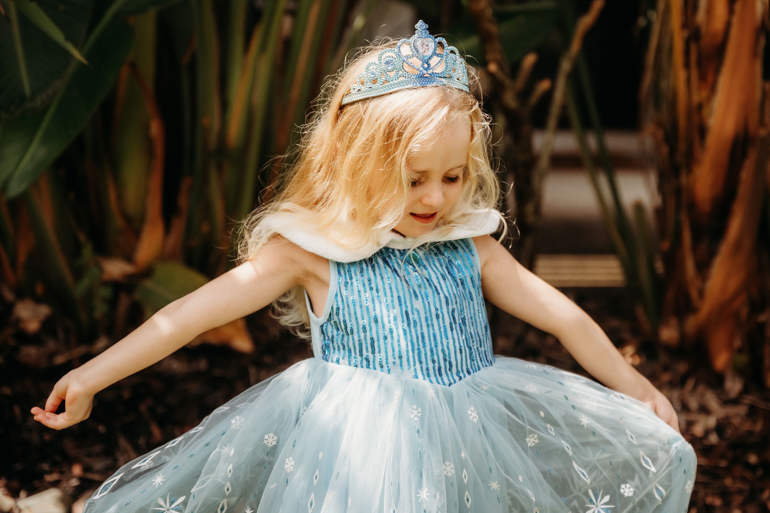 Young girl in blue sequin princess dress and tiara twirling outdoors among tropical plants.