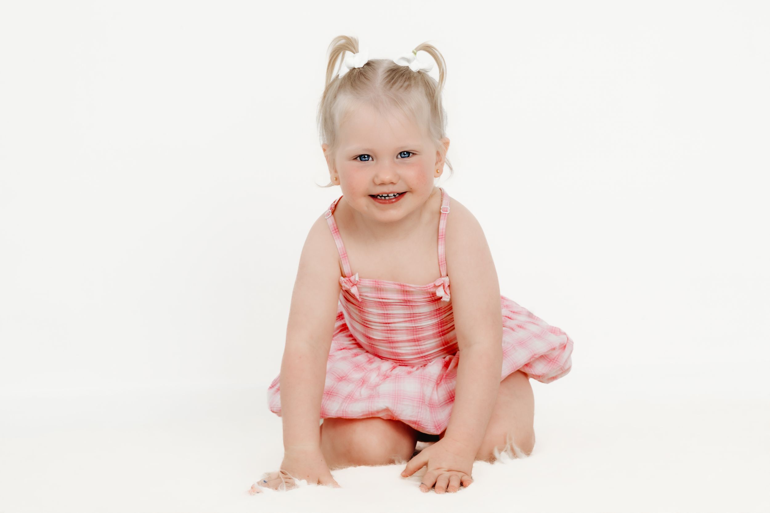 Smiling toddler girl in pink dress with pigtails sitting on white background.