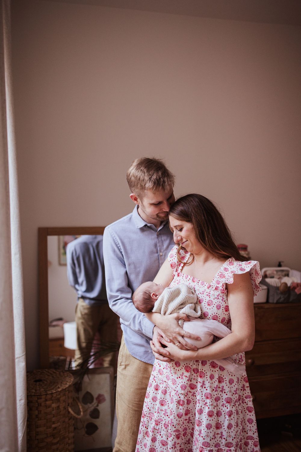 New parents tenderly gaze at their newborn baby, mother in floral dress, father in blue shirt, in a cozy nursery.