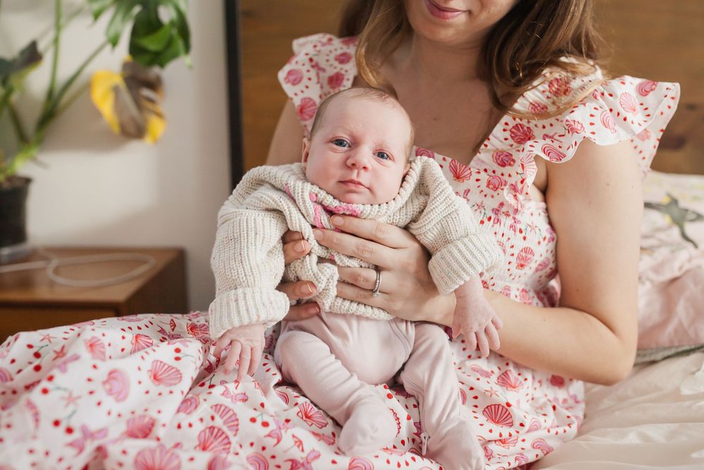 Mother in floral dress holds newborn baby in knit sweater while sitting on bed.