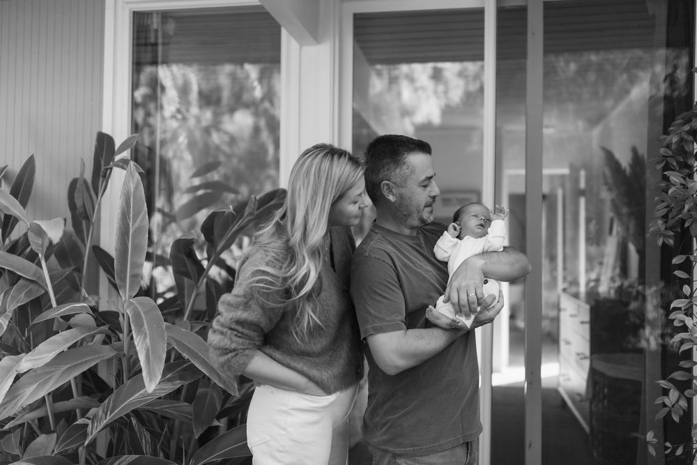 Black and white photo of a couple with a newborn baby standing outside their home surrounded by tropical plants.