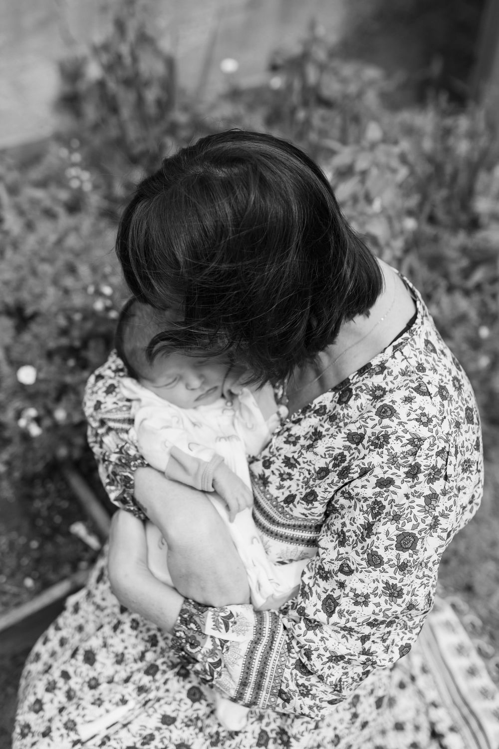 Black and white photo of a mother tenderly cradling her sleeping newborn baby outdoors among garden flowers.