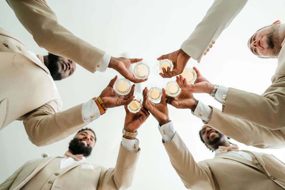 Group of men in cream suits toasting drinks in a circle, viewed from below at a wedding celebration.