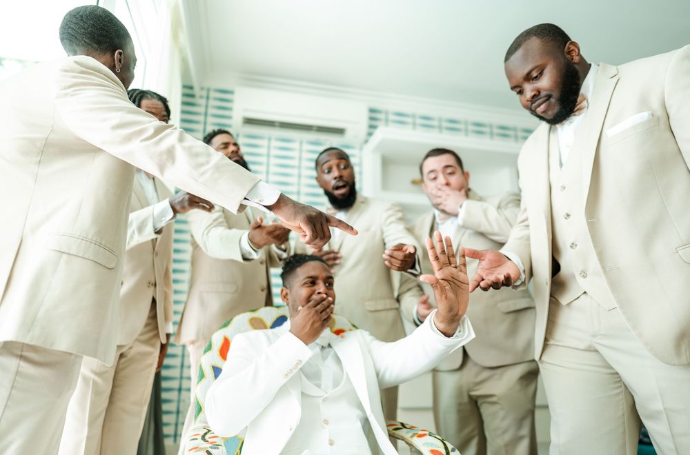 Groom in white suit with colorful accents seated as groomsmen in cream suits point toward him during wedding preparation.