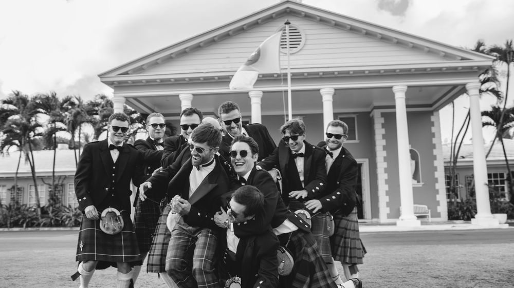 Groomsmen in kilts and suits celebrate with playful poses outside a white colonial building in a black and white photo.