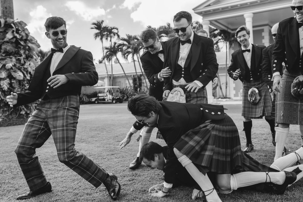 Men in kilts and suits laugh and play outdoors near palm trees in a candid black and white wedding photo.