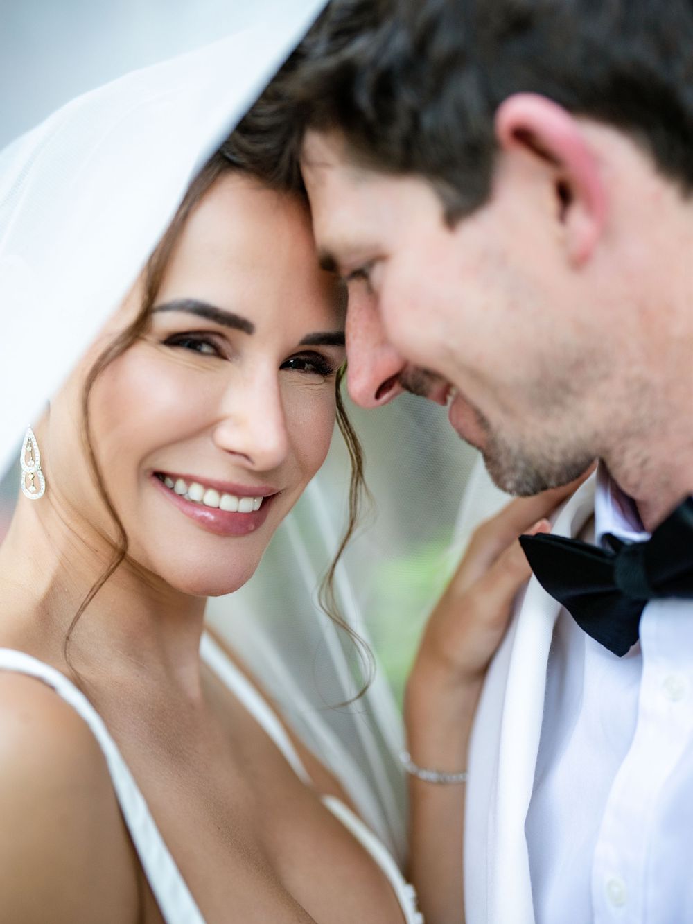 A romantic couple in white wedding attire share an intimate moment while smiling and embracing outdoors.